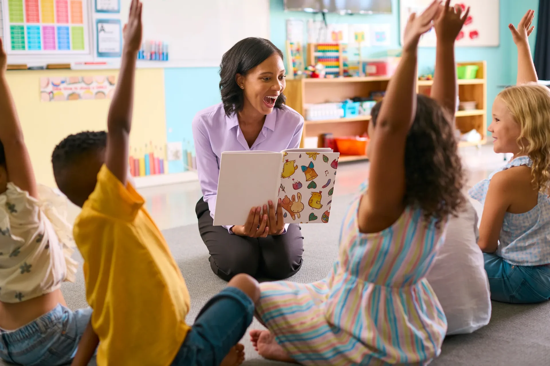 Teacher reading aloud to a group of engaged young students with raised hands